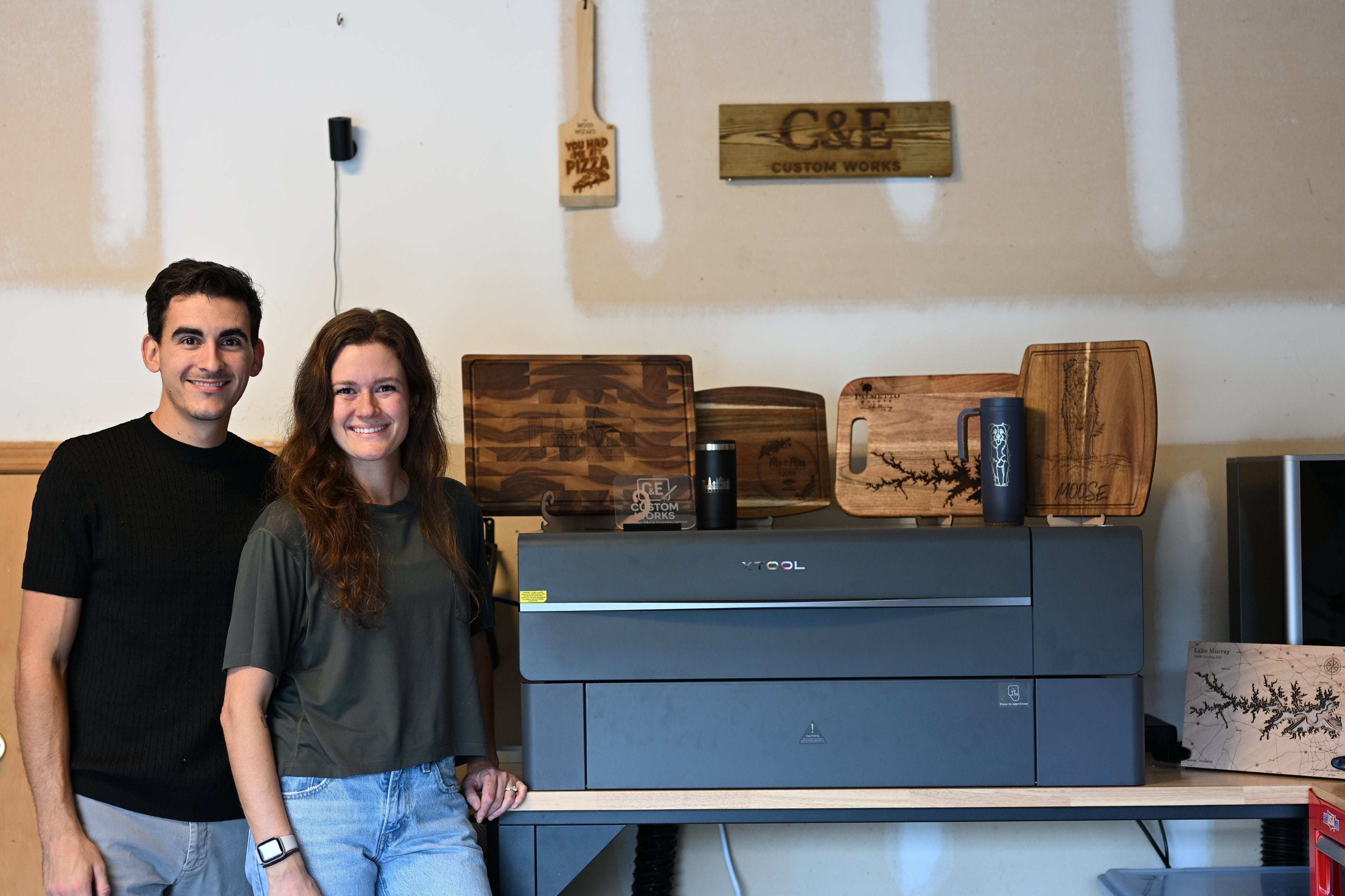 Two people standing in a workshop with wooden cutting boards and a laser engraving machine.